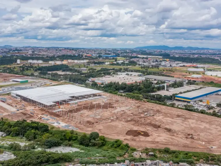 Vista aérea de grande terreno em terraplenagem para implantação de complexo logístico, com área urbana ao fundo.
