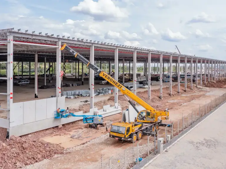 Vista aérea de guindaste erguendo estrutura metálica de galpão industrial em construção.