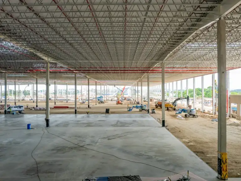 Fileira de colunas de concreto pré-moldado dentro de galpão industrial em construção, cobertura metálica ao fundo.