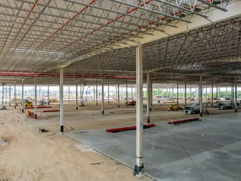 Fachada de vidro e estrutura de concreto de galpão industrial em construção, vegetação ao fundo.