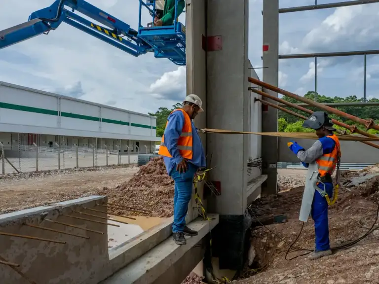 Dois profissionais em plataforma elevatória trabalham na estrutura metálica de um galpão industrial.