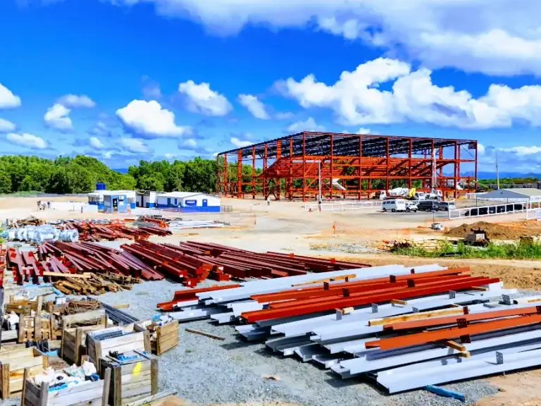 Estrutura metálica vermelha de edificação em construção, perfis de aço empilhados no canteiro.