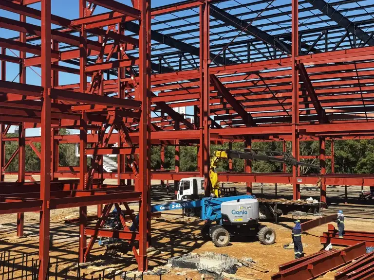 Estrutura metálica vermelha de edificação em construção, equipe trabalhando em plataforma elevatória.