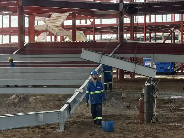 Estrutura metálica vermelha de galpão em construção, profissionais trabalhando na cobertura.