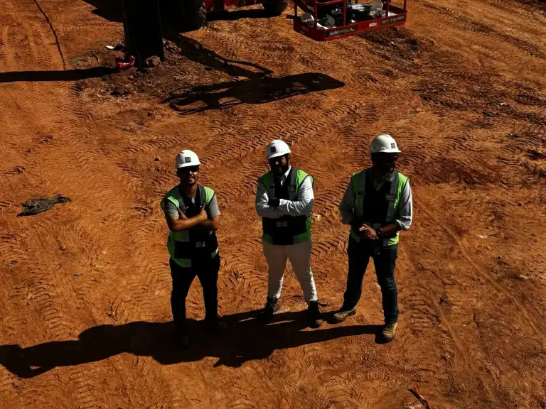 Três profissionais com capacete observam terreno em preparação para obra.