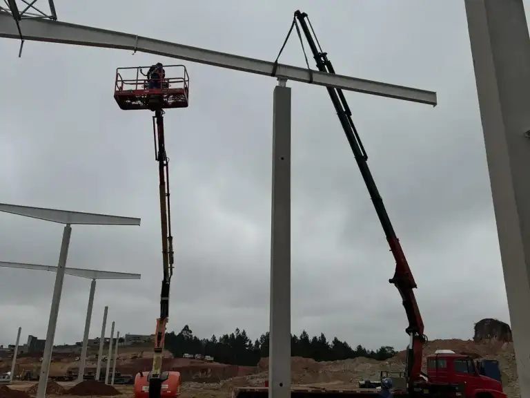 Guindaste e plataforma elevatória montando estrutura metálica de edificação sob céu azul.