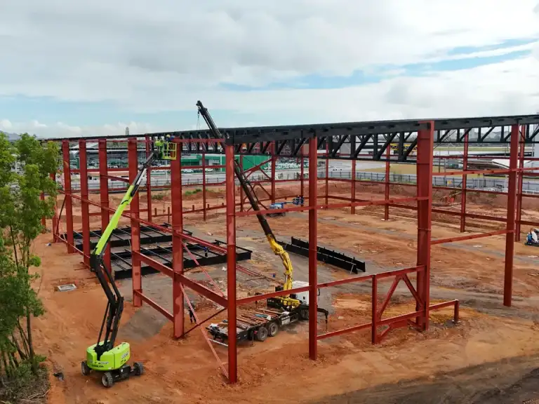 Estrutura metálica vermelha de edificação em construção, plataforma elevatória em operação.