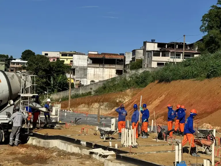 Equipe de profissionais e caminhão betoneira em fundação de edificação, área residencial ao fundo.
