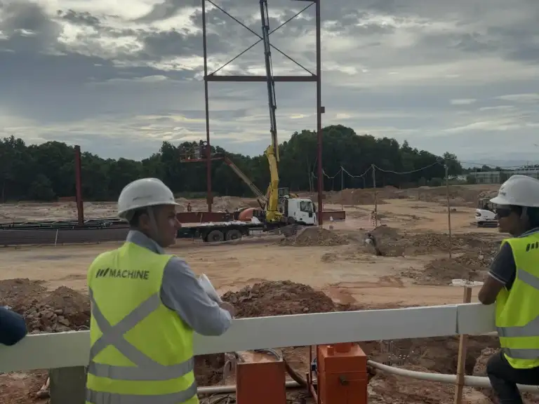 Profissional com colete de segurança acompanha estrutura metálica em montagem ao fundo.