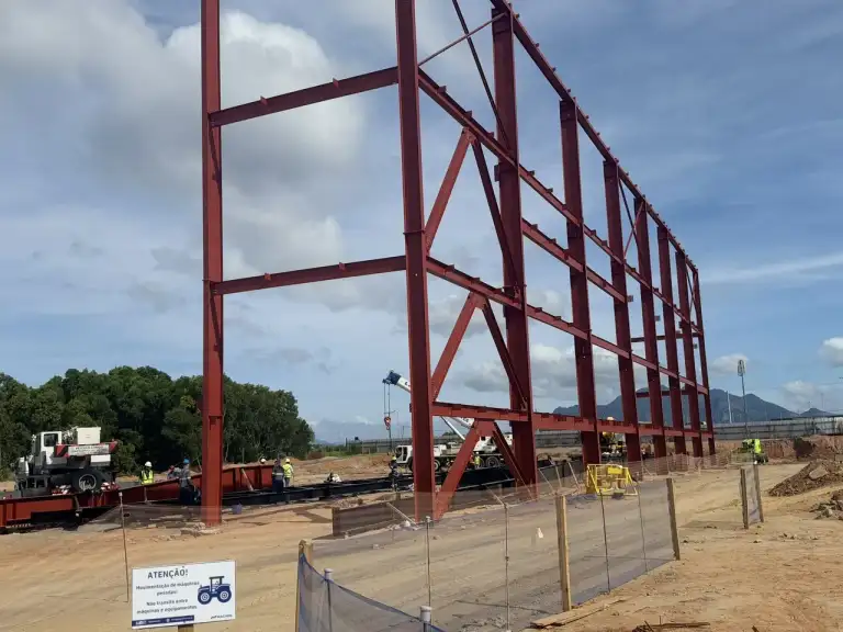 Estrutura metálica vermelha de edificação em construção, área urbana e montanhas ao fundo.
