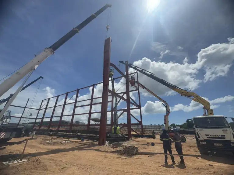 Guindaste erguendo pórtico de estrutura metálica vermelha contra céu azul com nuvens.